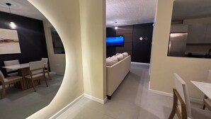 A modern living room with a sofa, a dining area with a table and chairs, and a kitchen area visible in the background. - Vive Luxe Condo , Pool & 24/7 Security near Airport (D'Abadie)