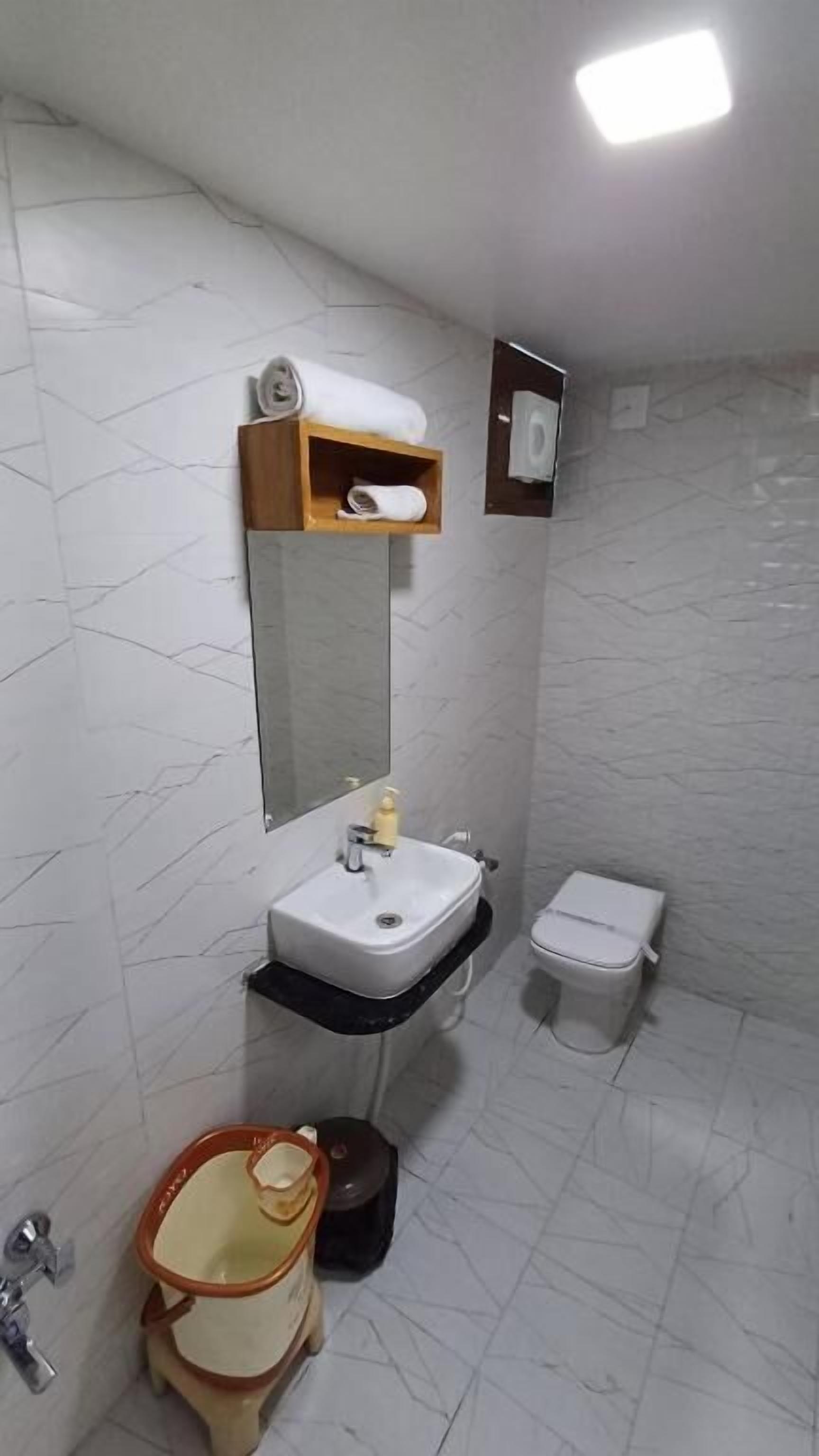 Une salle de bain avec un lavabo blanc, un miroir et des toilettes. On y trouve une étagère en bois avec des serviettes et un flacon de savon. Les murs sont recouverts de carreaux imitant le marbre.