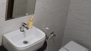A bathroom with a white sink, a mirror, a towel rack, a toilet, and a soap dispenser. - Hotel The Grand Jashi Residency (Patan)
