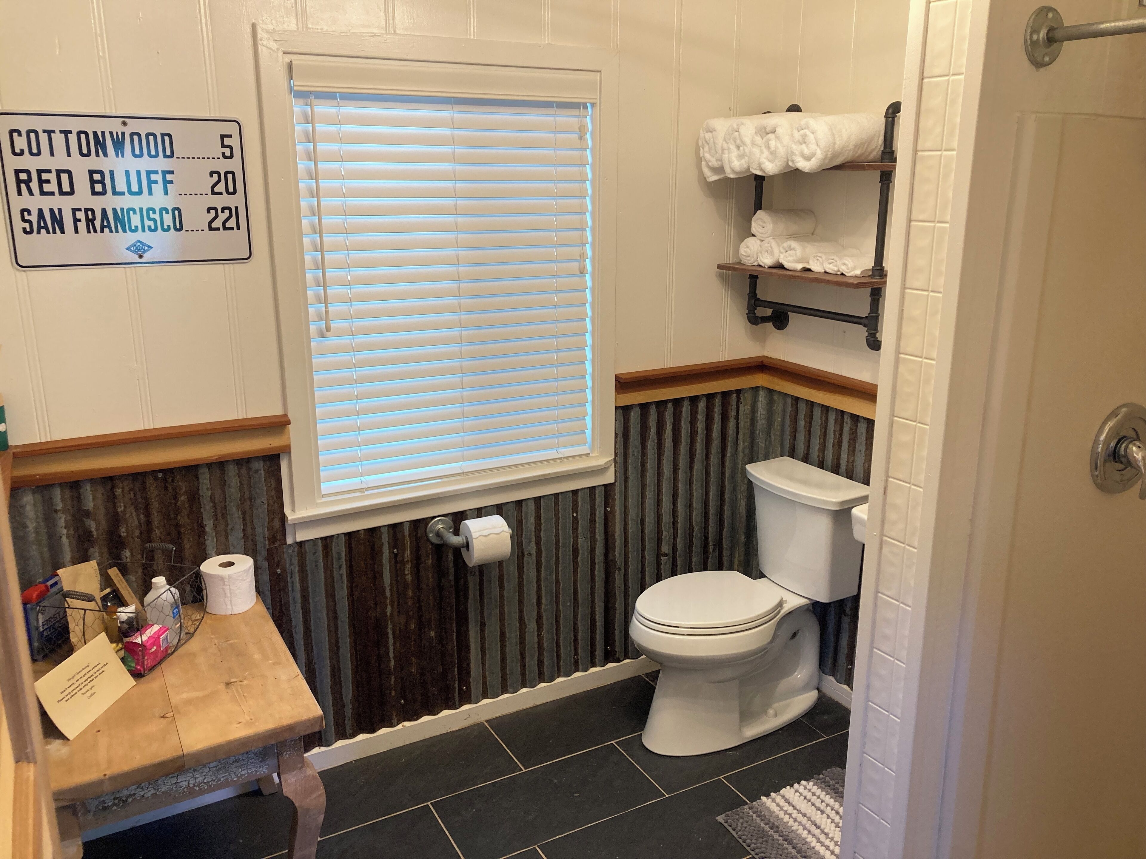 Une salle de bain équipée de toilettes, d’une fenêtre avec stores, d’un porte-serviettes et d’un panneau indiquant les numéros des locaux.