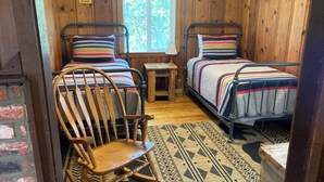 A cabin-style room with two beds, a rocking chair, a small table, and a window with a view of trees.