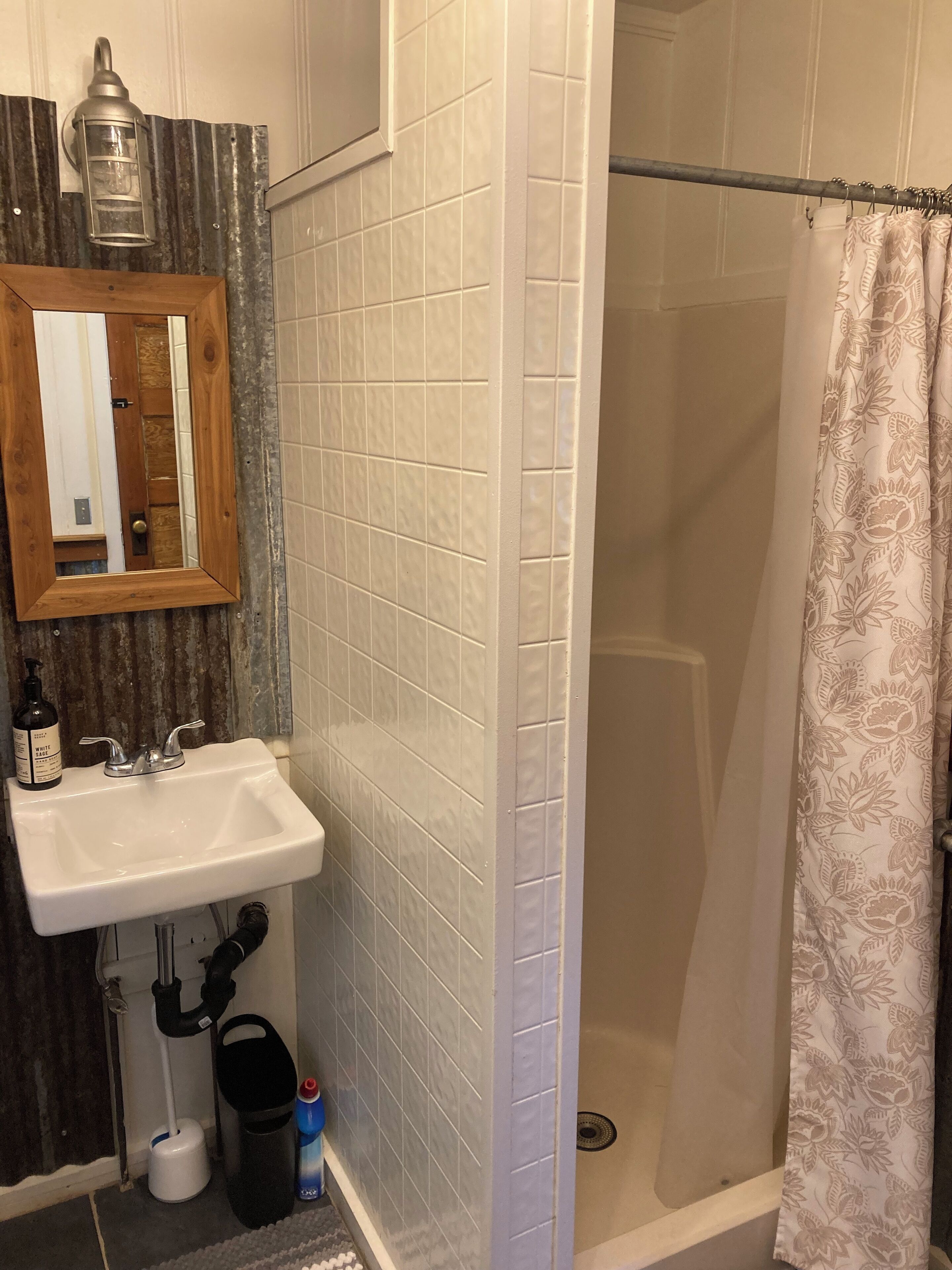 Une salle de bain avec un lavabo blanc, un miroir, une douche avec une porte en verre et un rideau de douche à motifs.