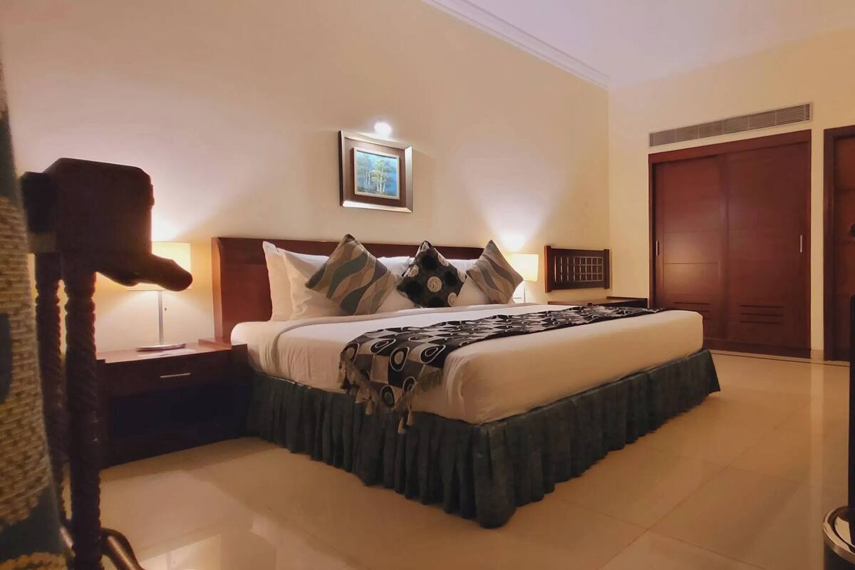 A hotel room with a bed, bedside table, lamp, framed picture, wardrobe, and a mirror.