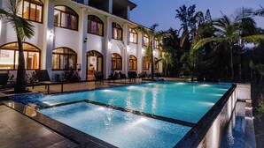 A large outdoor swimming pool surrounded by palm trees and a multi-story building with balconies. - Aeraki palms (Dahanu)