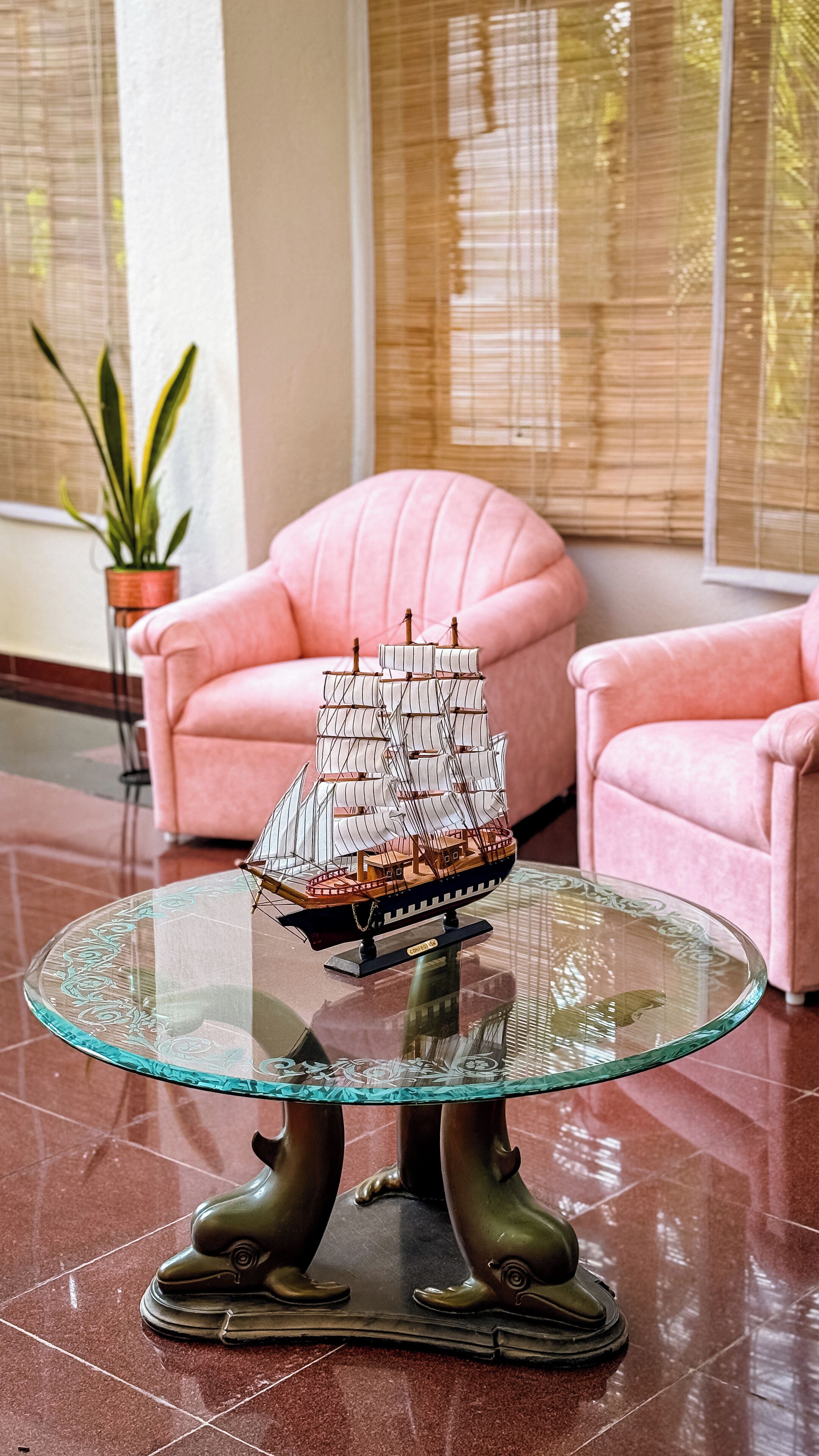 A model ship on a glass table in a room with pink chairs and a potted plant.