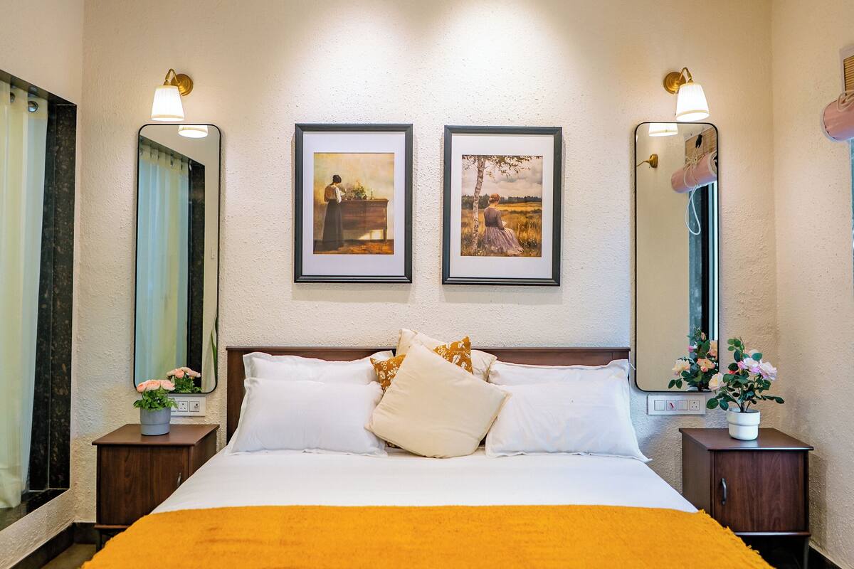 A bedroom with a bed, two framed pictures on the wall, a ceiling fan, and a nightstand with a plant.