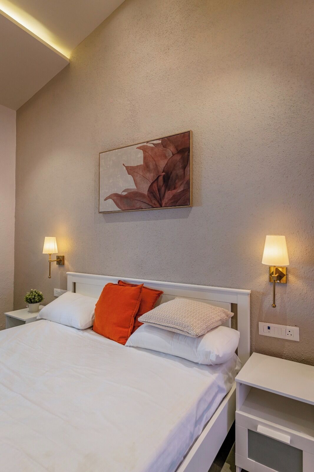 A neatly made bed with white and orange bedding, two bedside lamps, a wall-mounted artwork, and a small potted plant.