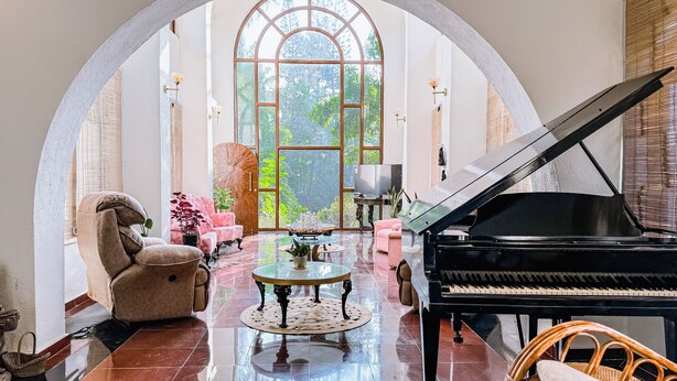 A spacious living room with a grand piano, comfortable seating, and a large arched window offering a view of lush greenery. - Aeraki palms (Dahanu)