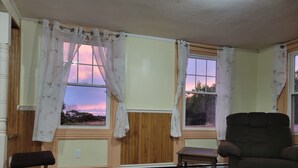 Habitación con suelo de madera, una silla de madera, una mesita y dos ventanas con cortinas.