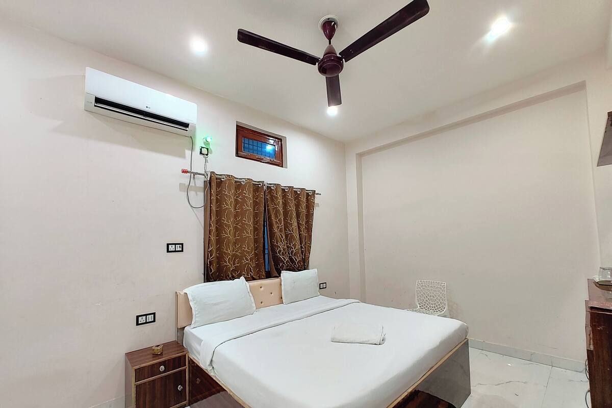 A bedroom with a bed, a ceiling fan, an air conditioner, a window with curtains, a bedside table, and a nightstand.