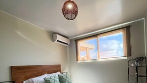 Een net opgemaakt bed met een houten hoofdbord, een groene plaid en opgerolde handdoeken. Er is een raam met uitzicht op de lucht en wolken, een airconditioning aan de muur en een plank met decoratieve objecten.