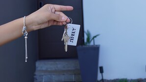Een hand die een sleutel en een sleutelkaart vasthoudt, met de naam 'STELLY' erop, voor een huis met een grijze deur en het huisnummer '18A'.