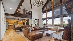 Een ruime woonkamer met een grote bruine leren bank, een eettafel en een stenen open haard. Achtergrond is een keuken met houten kastjes en een hoog plafond met zichtbare balken.