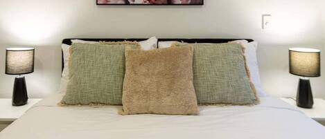 Ein ordentlich bezogenes Bett mit weißer Tagesdecke und drei Kissen in Grüntönen und Beige. Auf jeder Seite des Bettes stehen zwei Nachttischlampen.