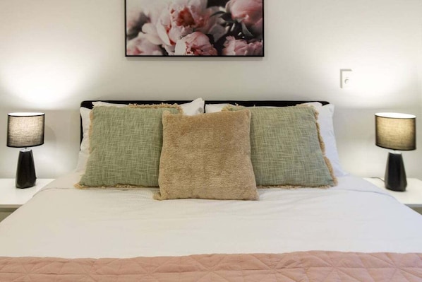 Ein ordentlich bezogenes Bett mit weißer Tagesdecke und drei Kissen in Grüntönen und Beige. Auf jeder Seite des Bettes stehen zwei Nachttischlampen.