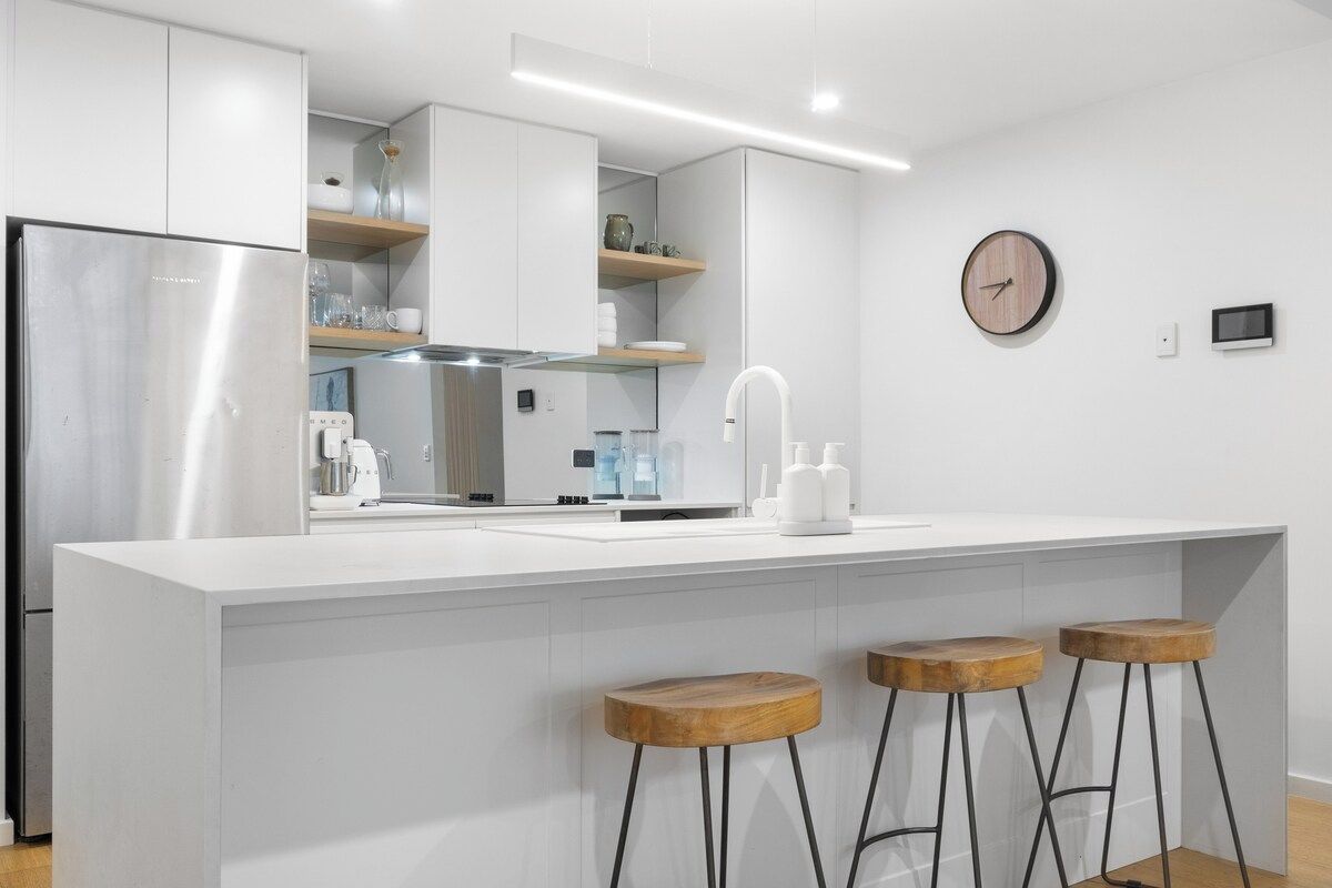 A modern kitchen with a stainless steel refrigerator, white cabinetry, a built-in oven, and a wooden bar stool seating area.