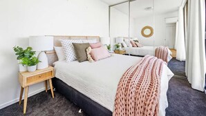Une chambre avec un grand miroir, un lit recouvert d’une couette blanche, une table de chevet avec une lampe et une plante en pot.