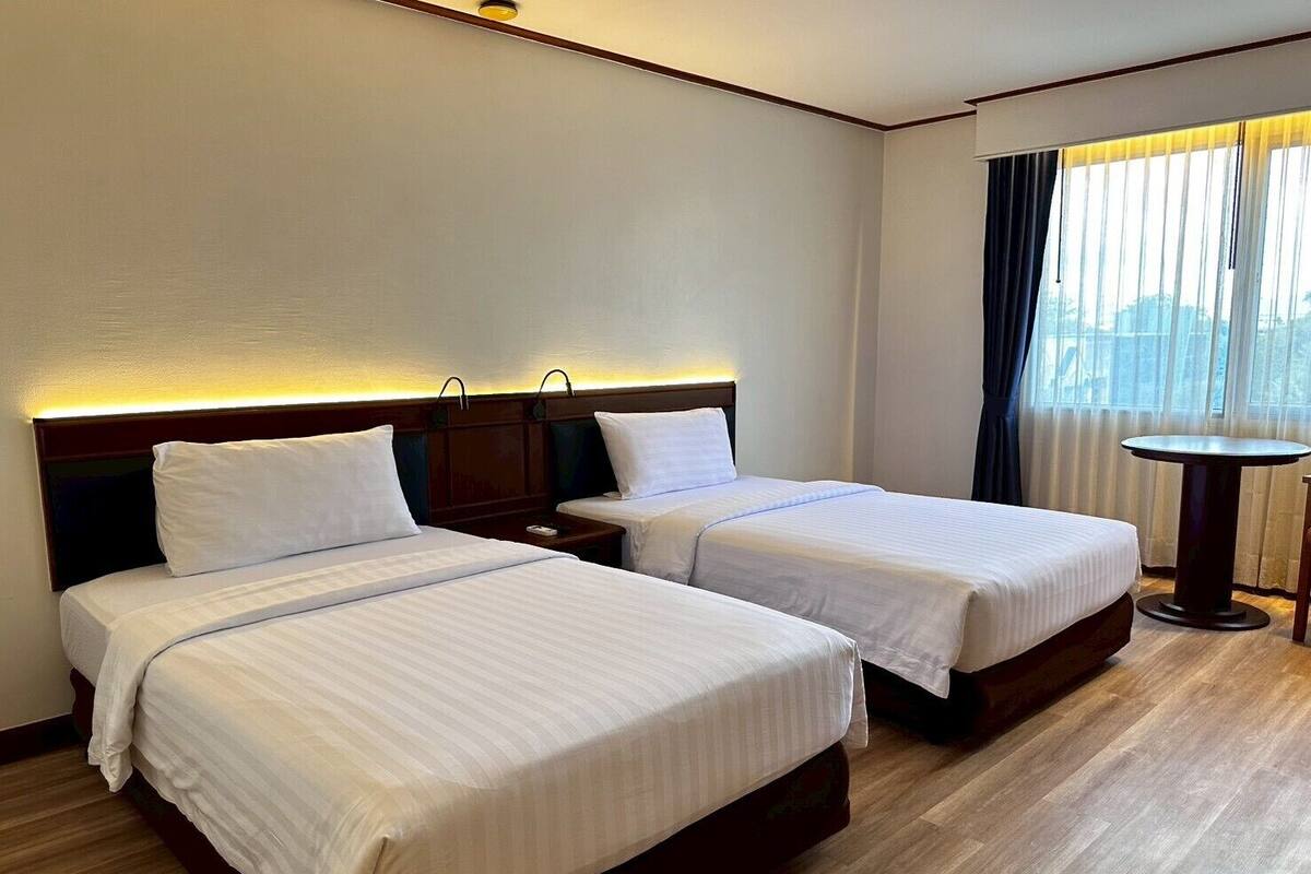 A hotel room with two beds, a small table, and a window with curtains.