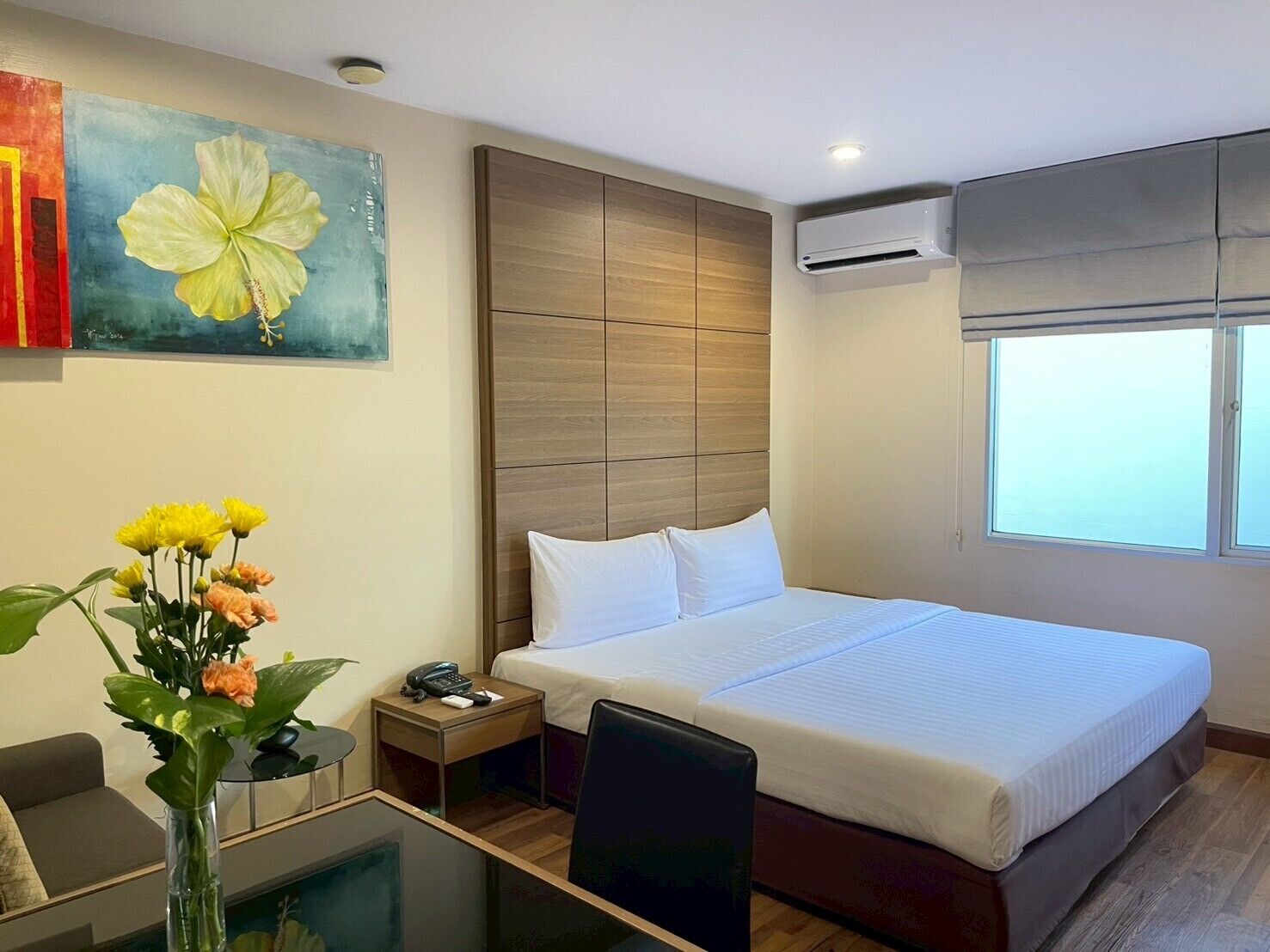 Habitación de hotel con una cama grande, cabecera de madera, escritorio con silla, mesita de noche con teléfono y ventana con persianas.