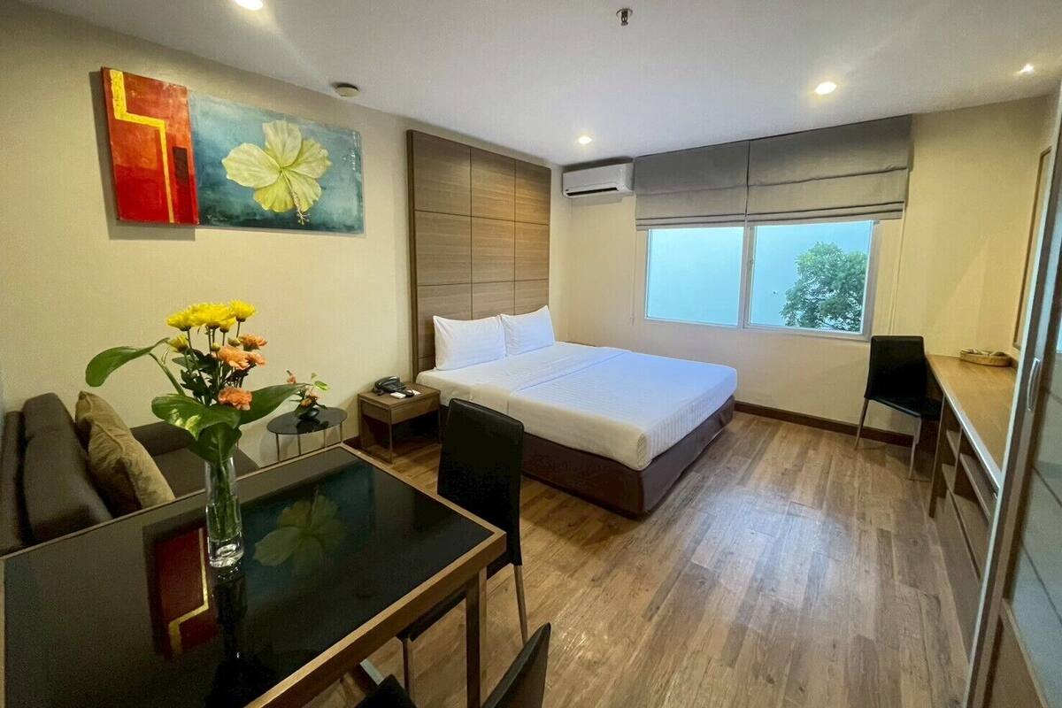 A hotel room with a bed, a desk, a chair, a small table, a vase with flowers, and a painting on the wall.