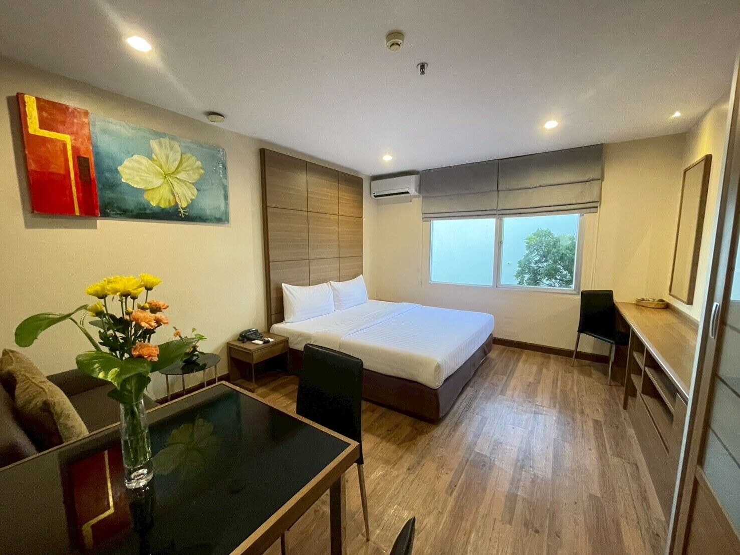 A hotel room with a large bed, a desk, a chair, a small table with a vase of flowers, and a window with a view of trees.