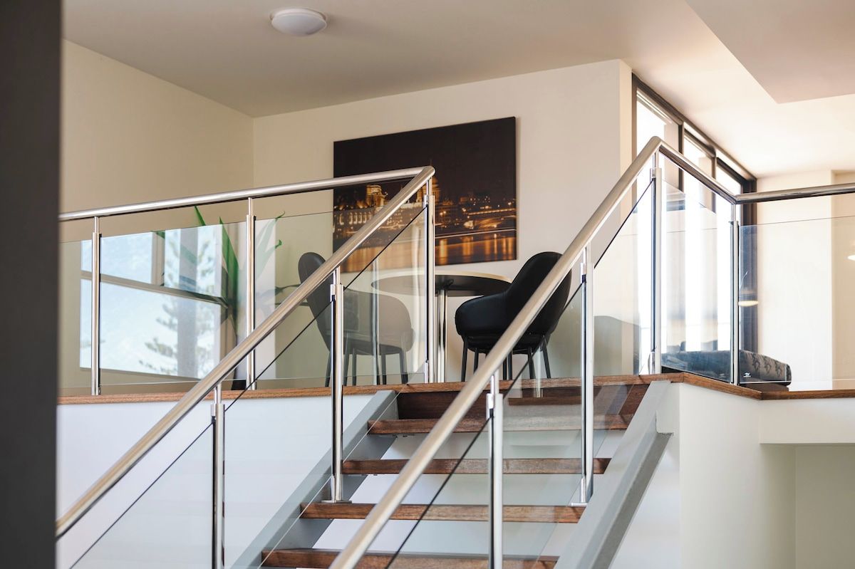 Una escalera moderna con barandillas de cristal y peldaños de madera que conduce a un comedor con mesa y sillas.