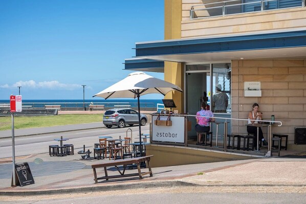 An outdoor seating area with tables and chairs, a sign for 'estabar', and a view of the ocean.