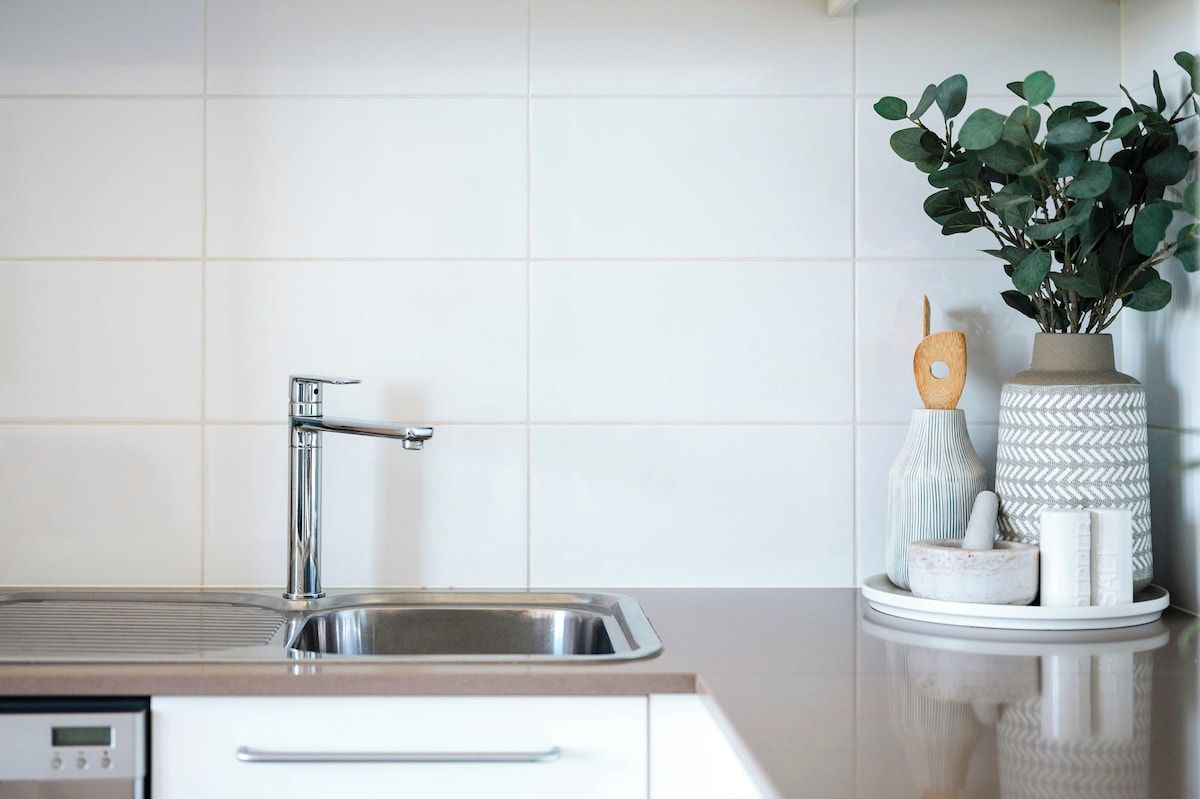 Una cocina con un lavamanos de acero inoxidable, una llave moderna y un salpicadero blanco de azulejos.