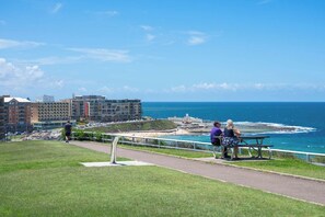 Vista costera con un paseo pavimentado, un banco y una zona ajardinada.