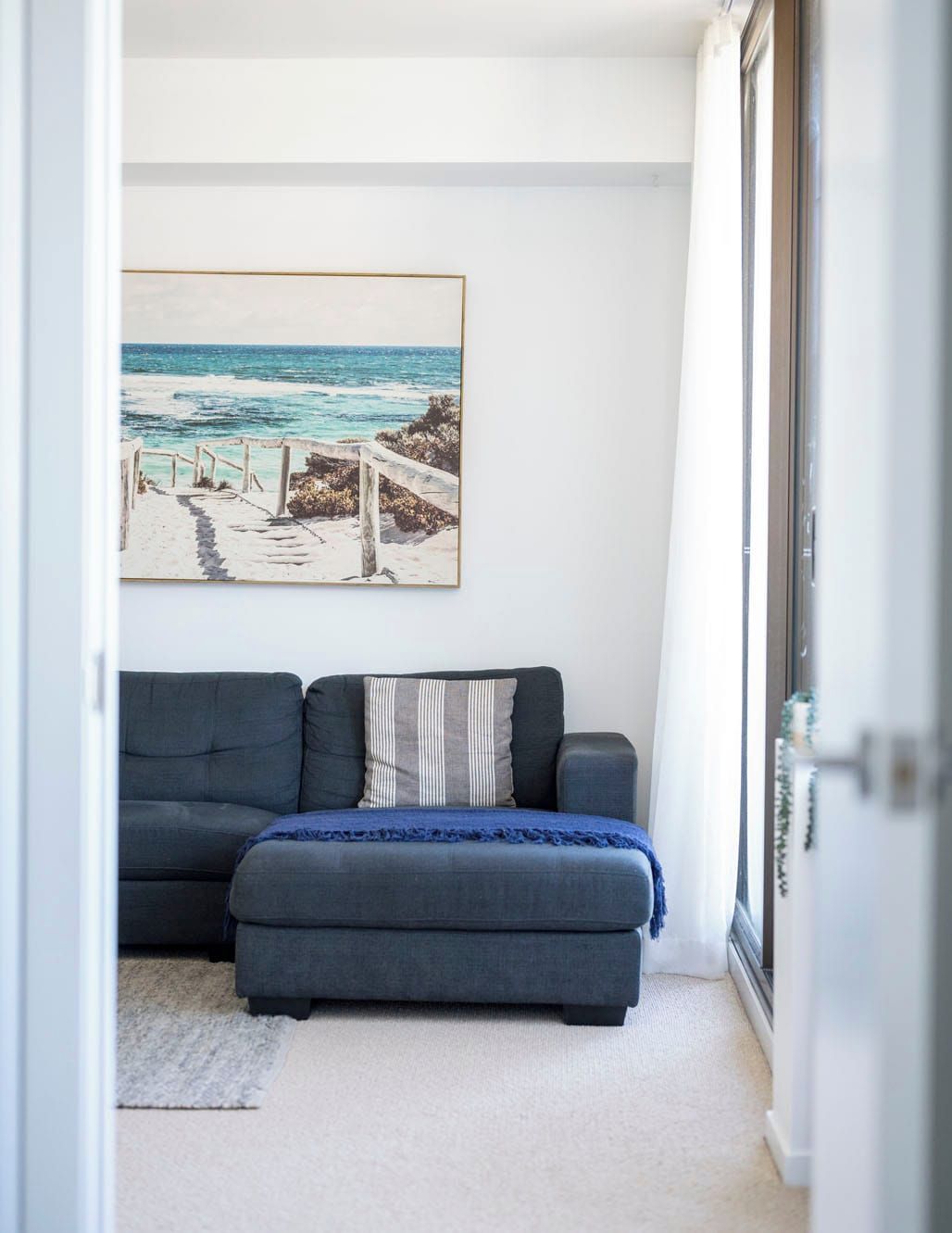 Una habitación con un sofá gris, un cuadro enmarcado de playa y vistas a través de un ventanal.