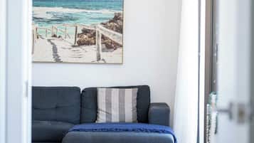 Una habitación con un sofá gris, un cuadro enmarcado de playa y vistas a través de un ventanal.