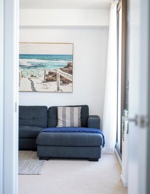 Una habitación con un sofá gris, un cuadro enmarcado de playa y vistas a través de un ventanal.