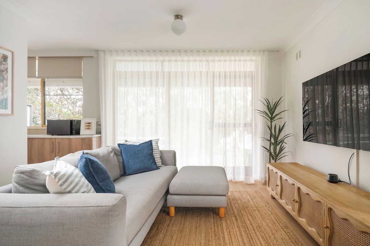 A modern living room with a grey sectional sofa, a wooden console table, and a large window with sheer curtains.