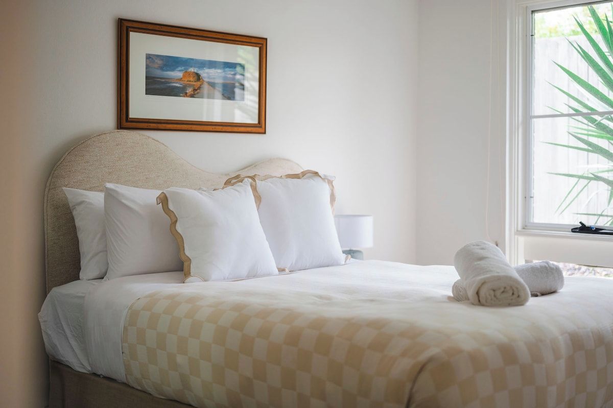 A neatly made bed with a checkered bedspread, a framed picture on the wall, and a window with a plant outside.