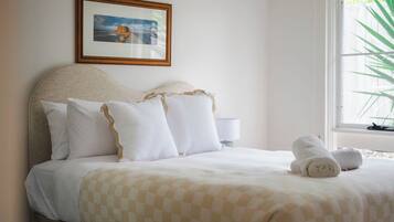 A neatly made bed with a checkered bedspread, a framed picture on the wall, and a window with a plant outside.