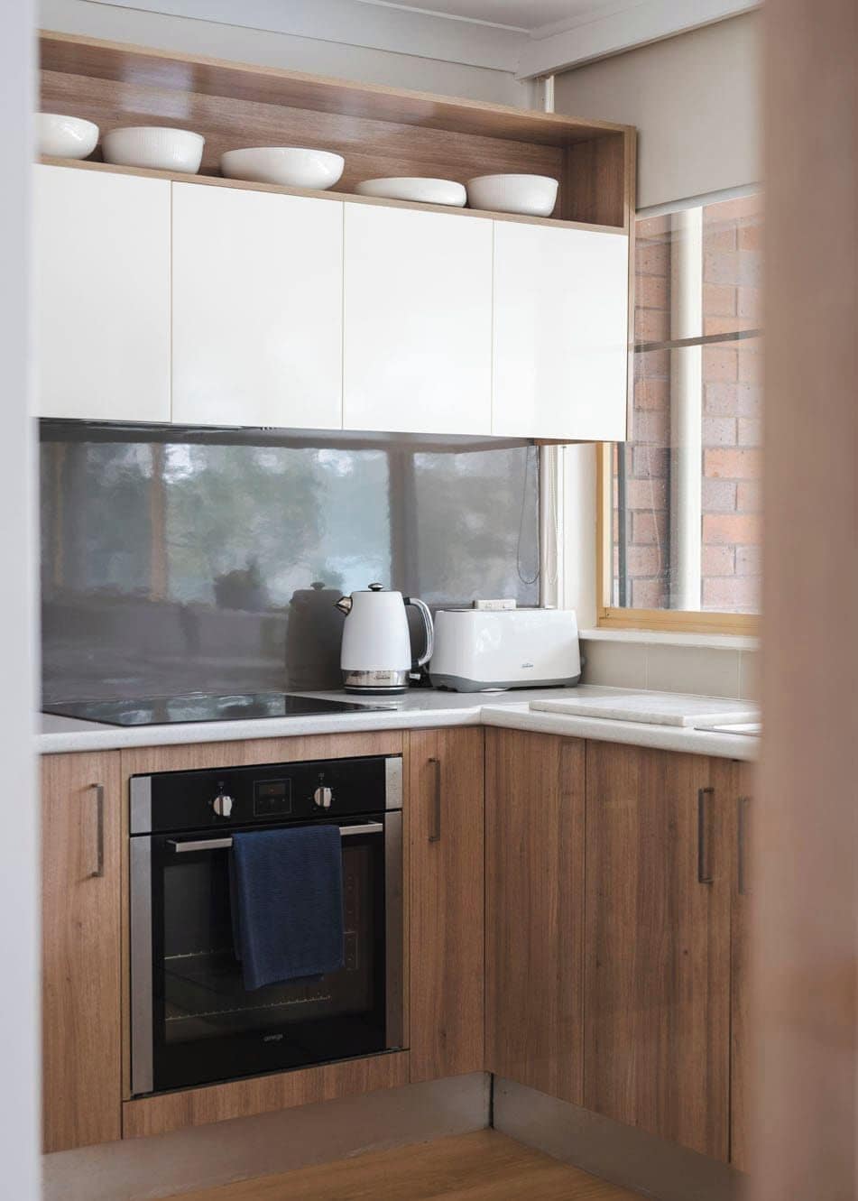 A compact kitchen with white cabinets, a built-in oven, and a window with a view of a brick wall.