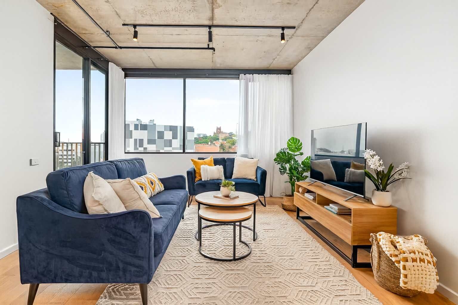 A modern living room with a blue sofa, a smaller blue armchair, a round coffee table, a wooden TV stand, and a large window with a city view.