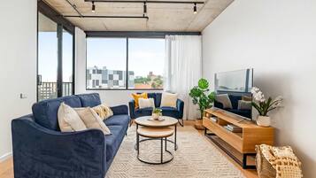 A modern living room with a blue sofa, a smaller blue armchair, a round coffee table, a wooden TV stand, and a large window with a city view.