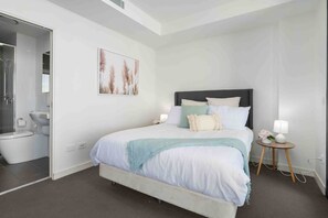 A modern bedroom with a large bed, a bedside table with a lamp, and a bathroom visible through an open door.
