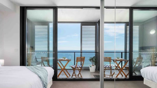 Habitación de hotel con dos camas, un balcón con vista al mar y zona de comedor con sillas y una mesa.
