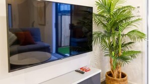 A flat-screen TV mounted on a white wall, a potted plant beside it, and a remote control on the TV stand.