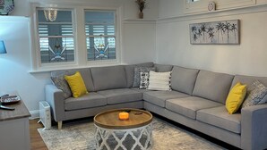 A living room with a grey sofa, a round coffee table, a chandelier, and a painting of palm trees.