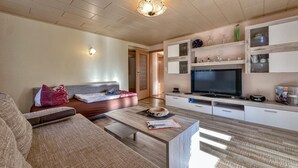 Een moderne woonkamer met een bank, salontafel en televisie. Er is een houten plafond en een deur naar een andere kamer.