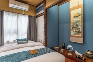 Habitación de hotel con cama, aire acondicionado de pared, pintura de montaña, una mesita con un florero, un espejo y una figurita decorativa.