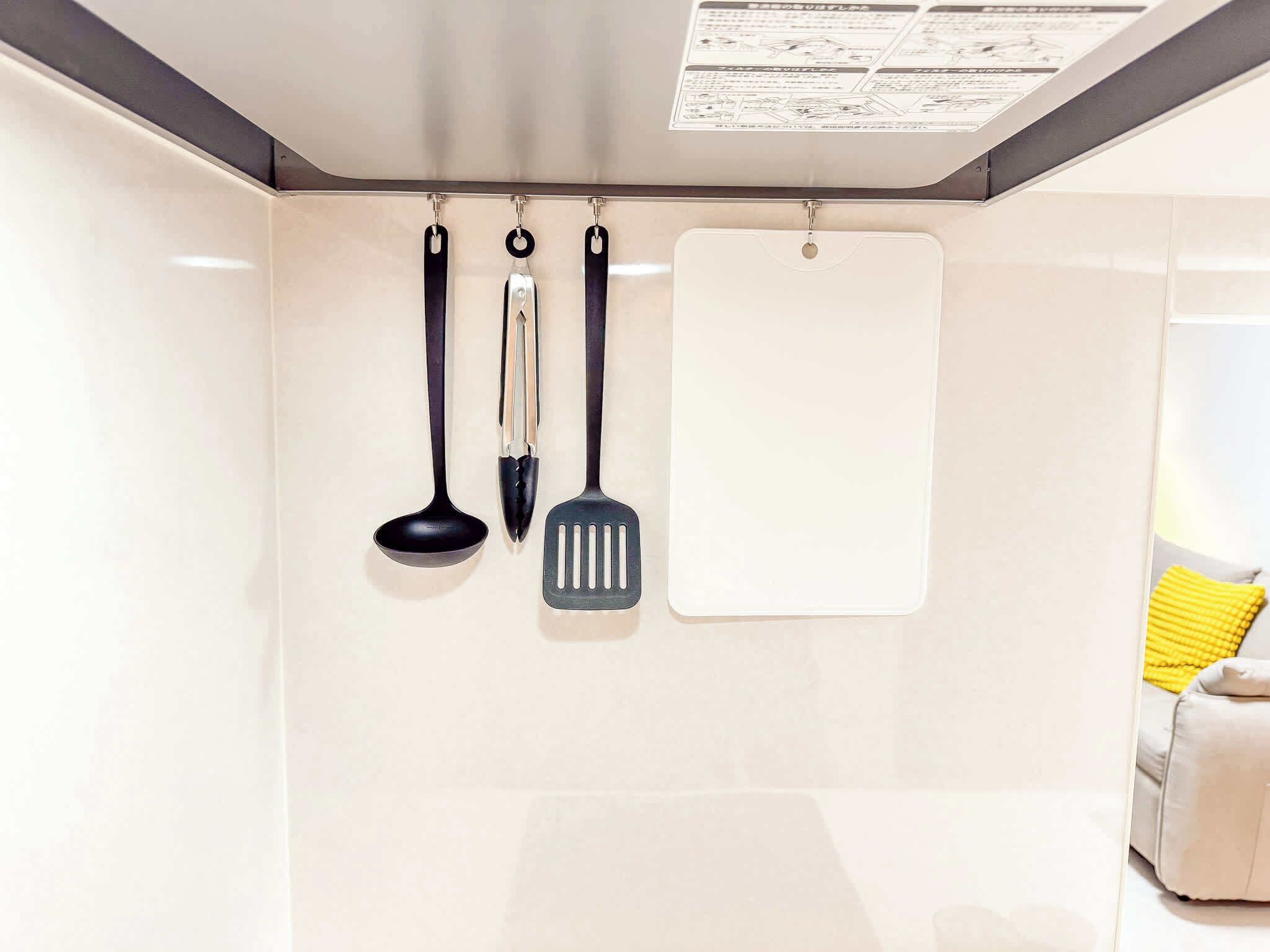 A kitchen with three utensils hanging on a wall, a whiteboard, and a yellow cushion on a sofa.