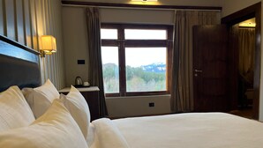 A hotel room with a large bed, a desk with a lamp, a window with a view of mountains, and a door. - Hotel Raks  (Leh)