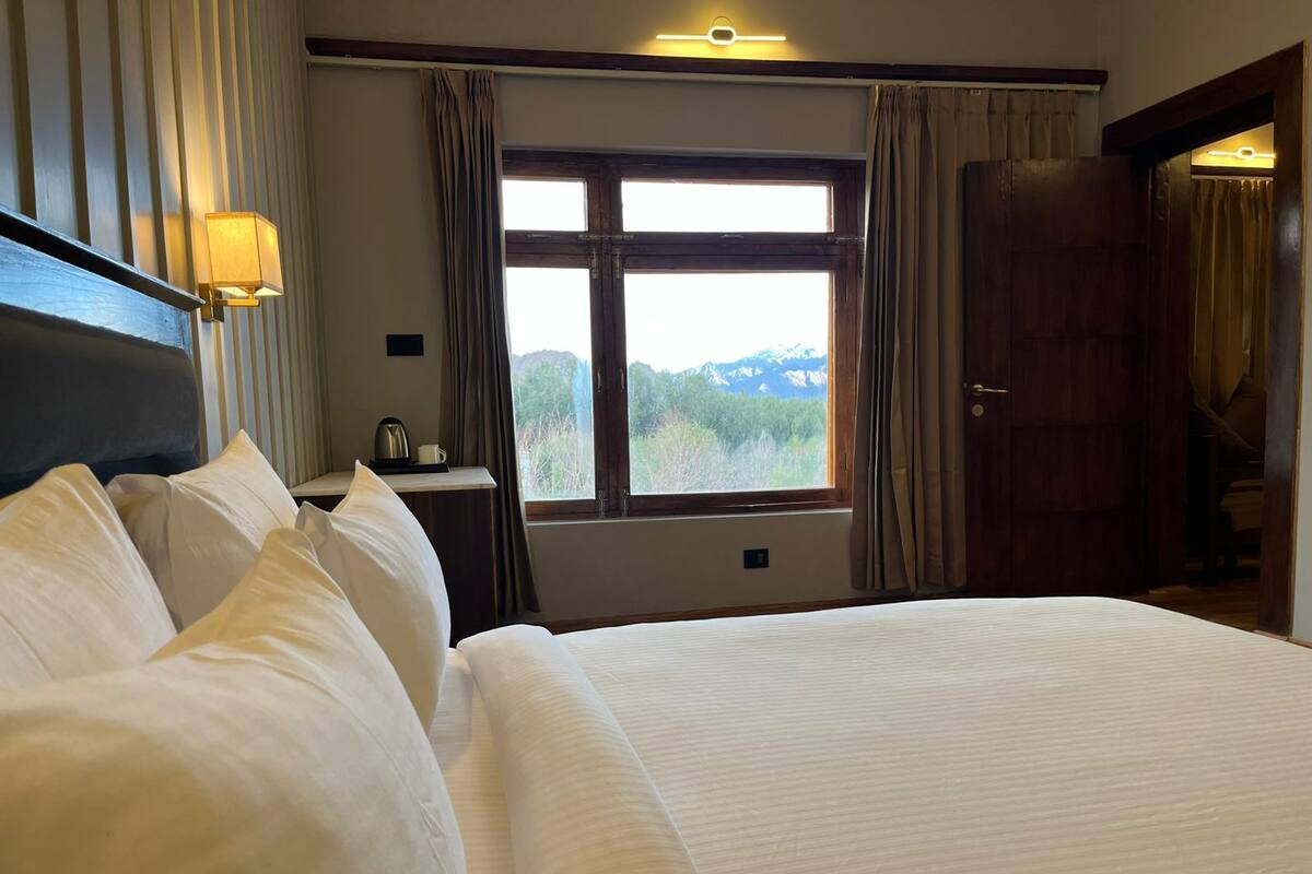 A hotel room with a large bed, a desk with a lamp, a window with a view of mountains, and a door.