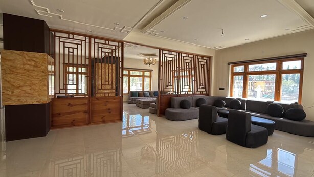 A spacious living area with a modern sofa set, a wooden partition, and large windows providing natural light. - Hotel Raks  (Leh)