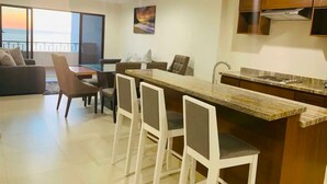 A modern kitchen with a bar area, dining chairs, and a large window overlooking a balcony. - Luxury apartment with ocean view in the golden zone (Mazatlán)
