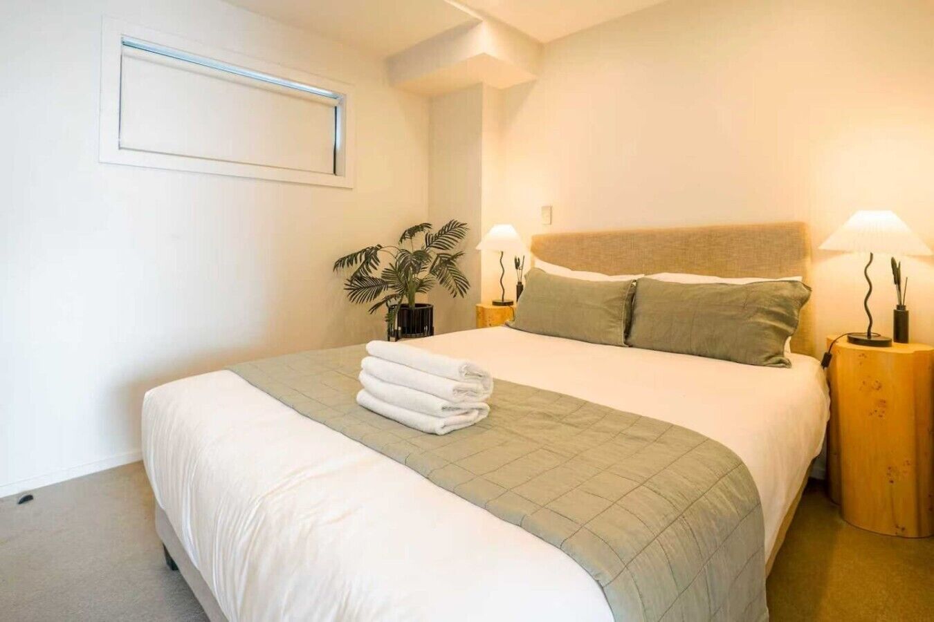 A neatly made bed with a green bedspread and white linens, two bedside lamps, a potted plant, and a wall-mounted light fixture.